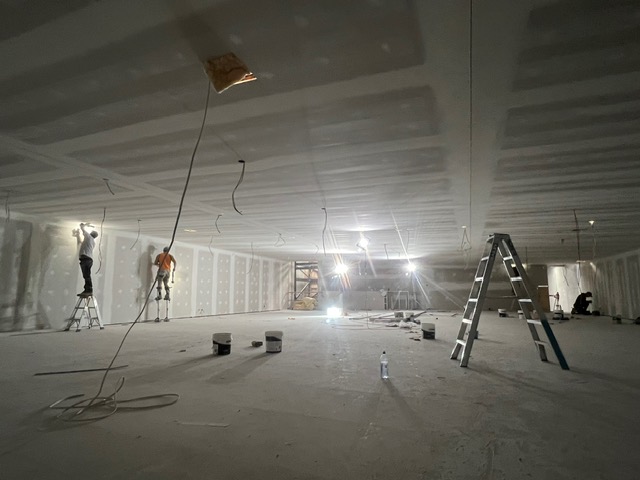 Workers on stilts and ladders setting plasterboard walls in a large open commercial space